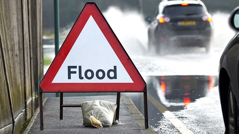 Image of a flood warning sign, in a red triangle on a road where cars are driving through water.