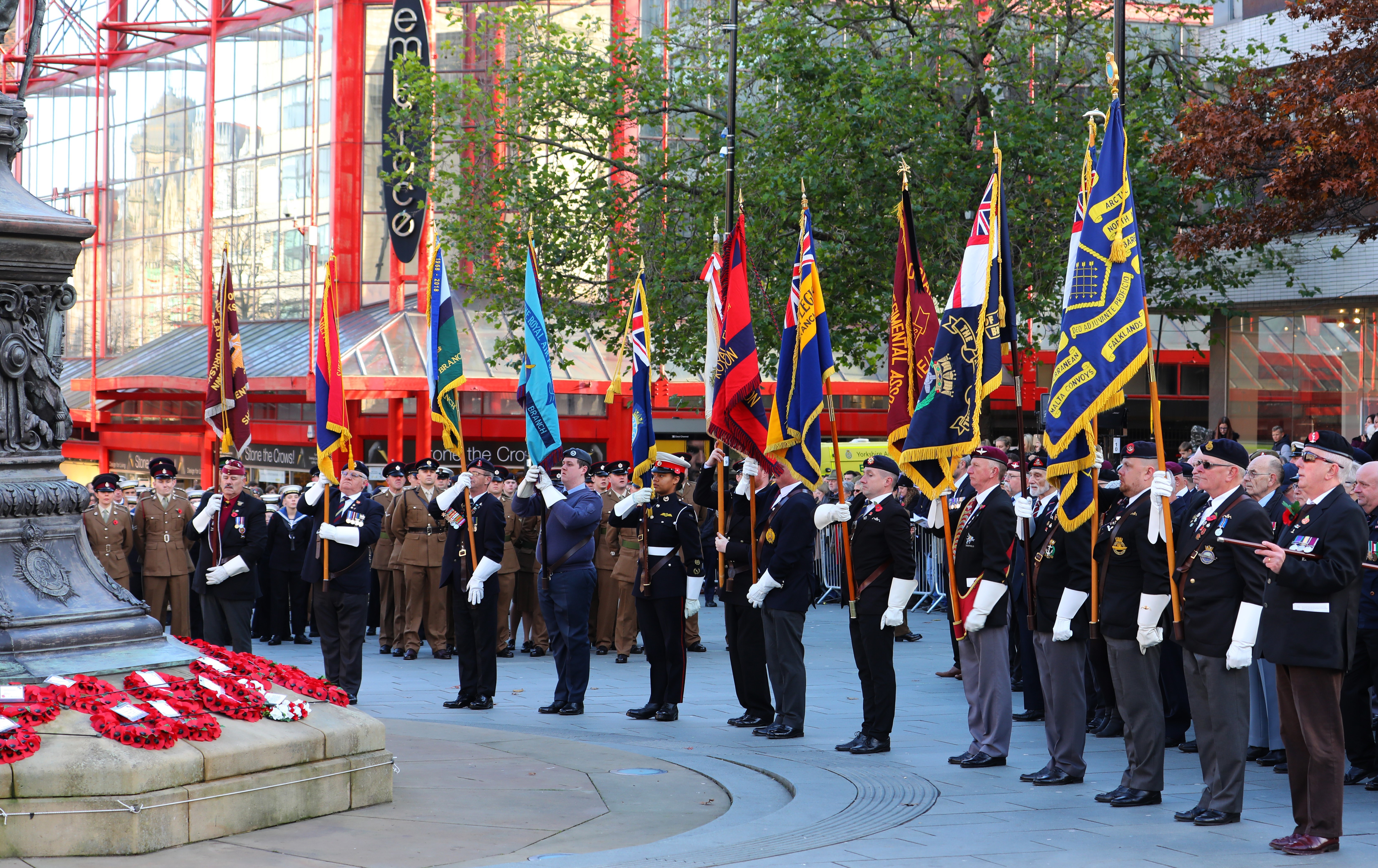 Men dressed in military uniform stand in a line holding flags, poppy wreaths lay in front of a monument