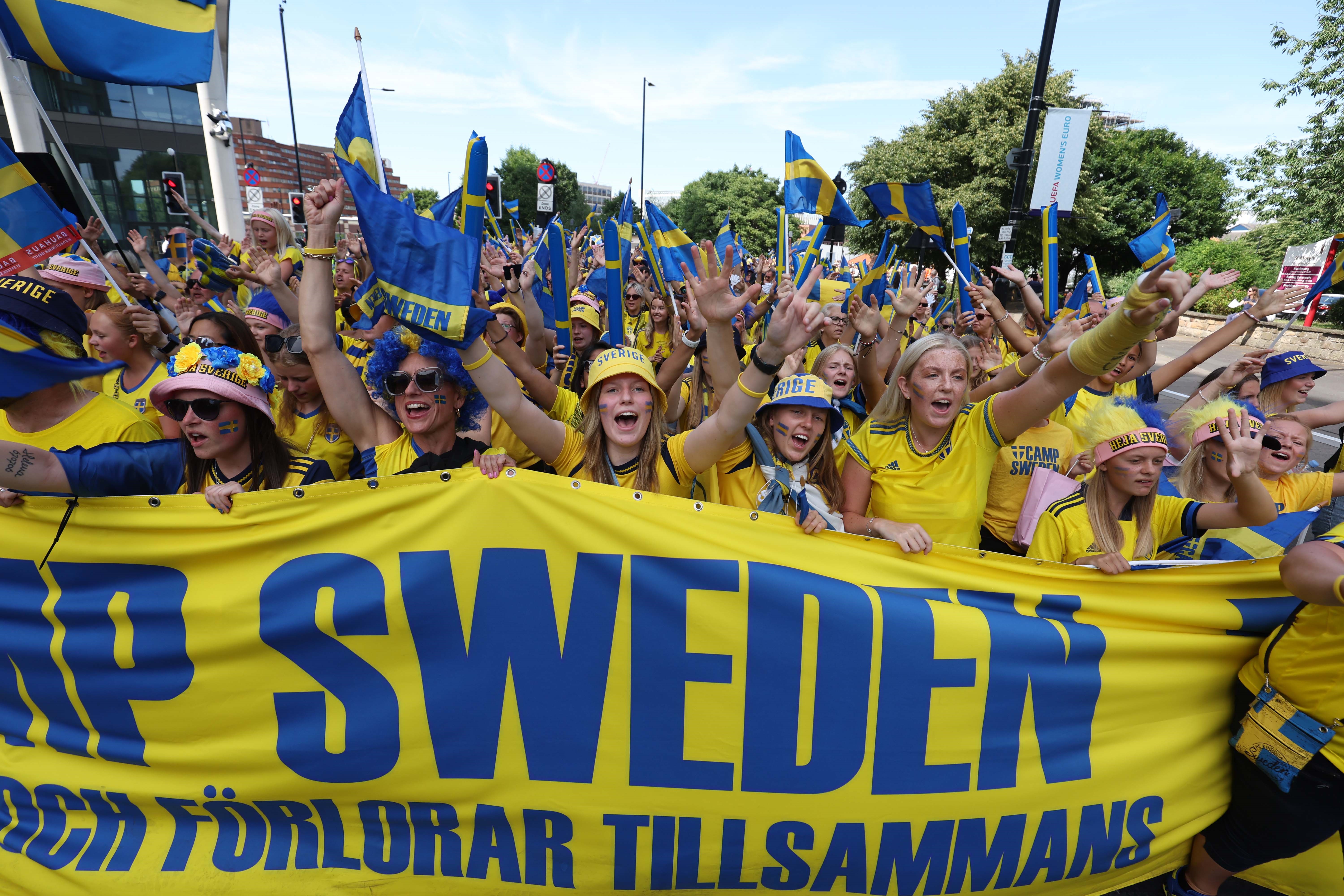 A large group of people dressed in yellow hold a large banner that say 'camp Sweden'