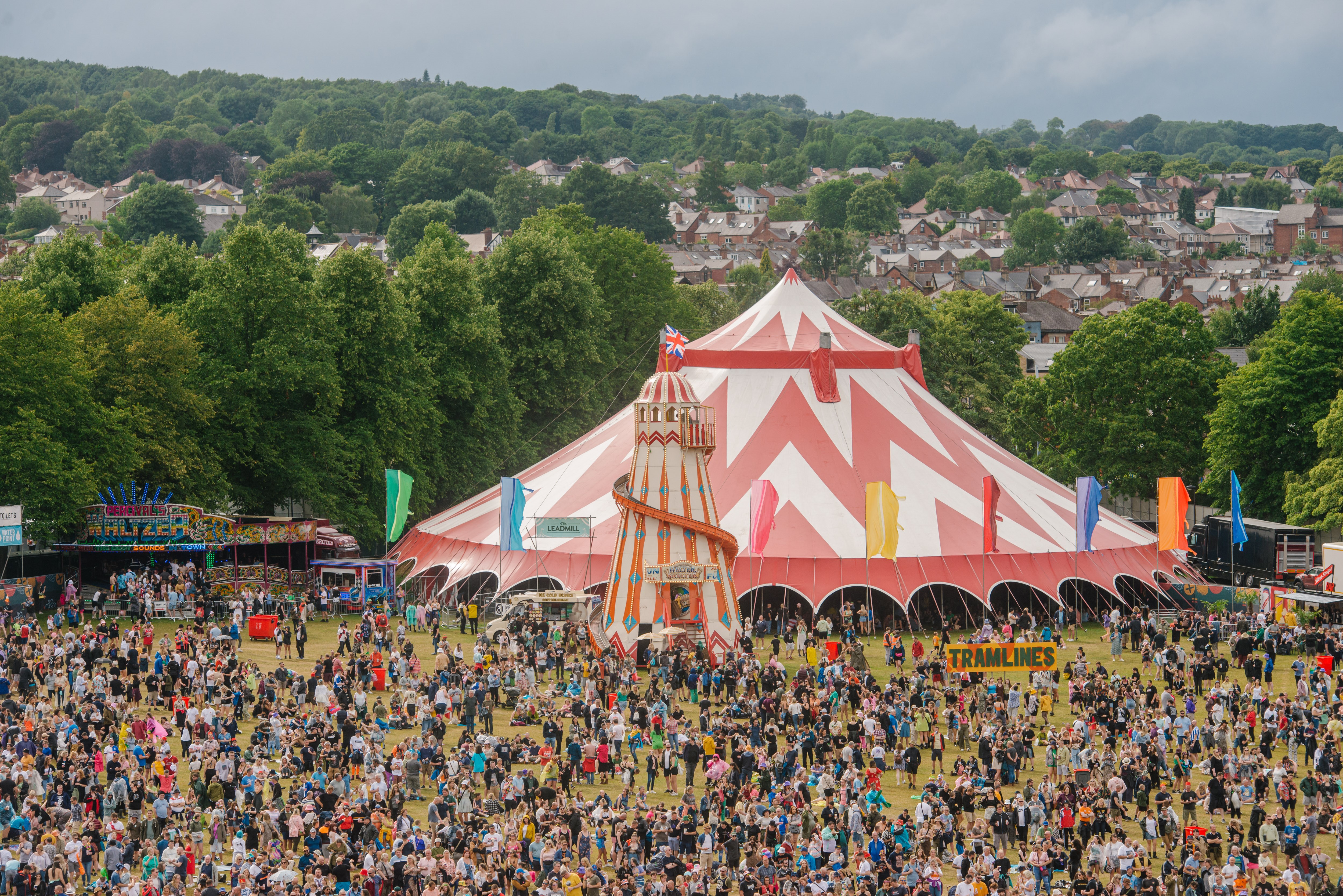 A huge crowd of people stand around a large festival tent, trees are in the background