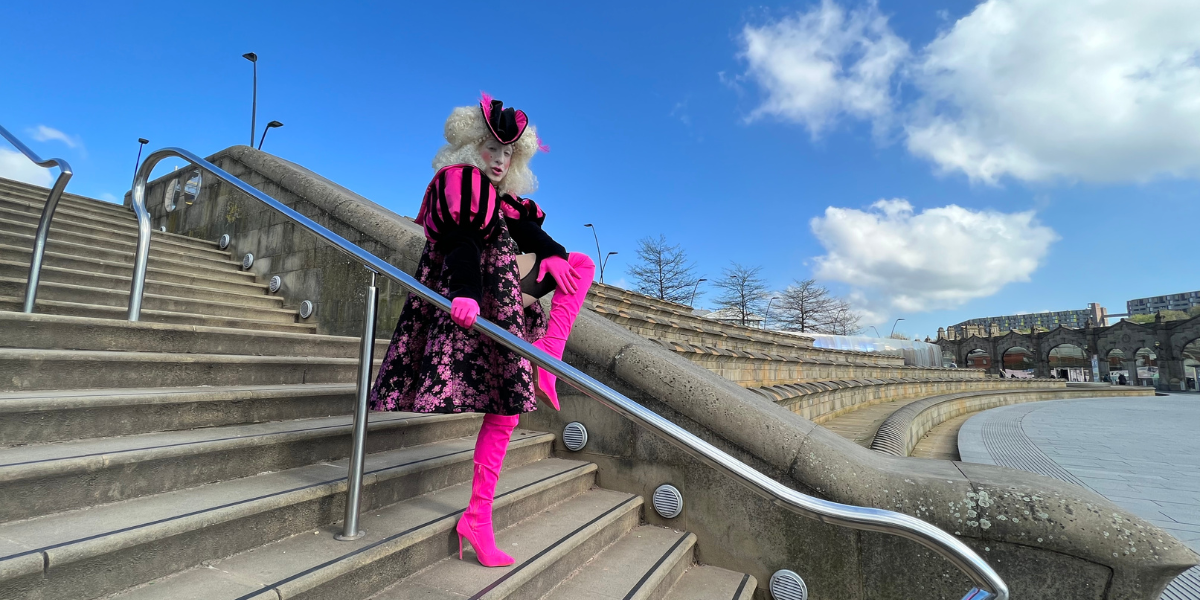 Jamie is wearing a pink and white outfit and a white curly wig. He is standing on some steps looking down at the camera and posing.