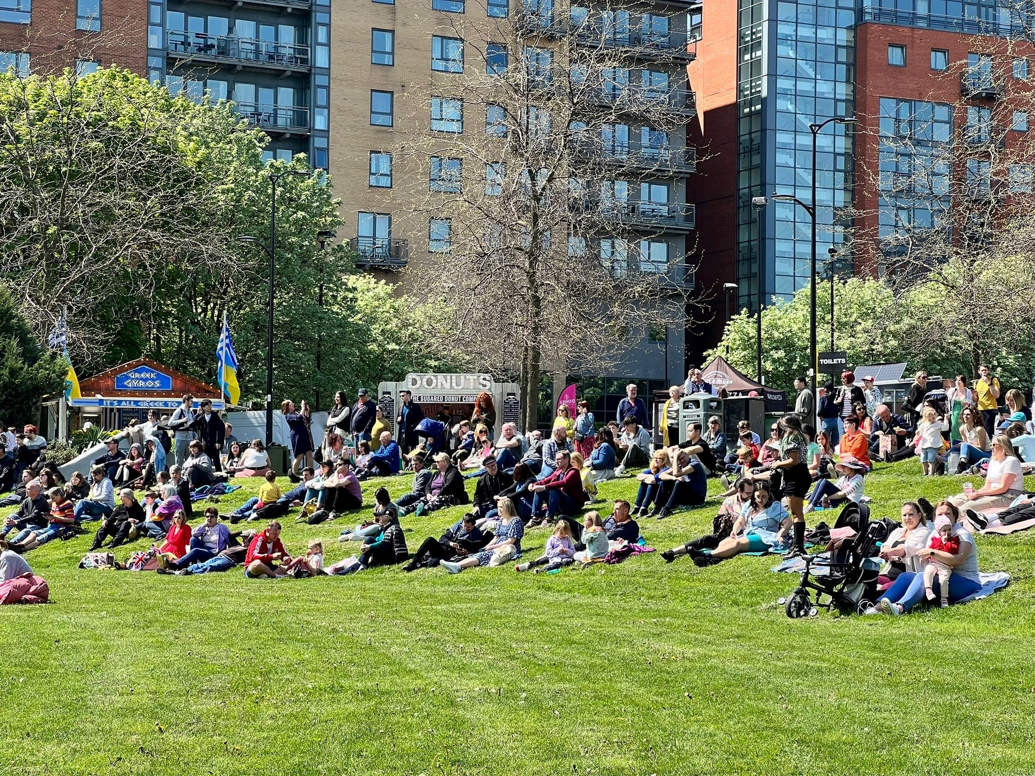 People sit on Devonshire Green in the sunshine watching acts perform on stage.