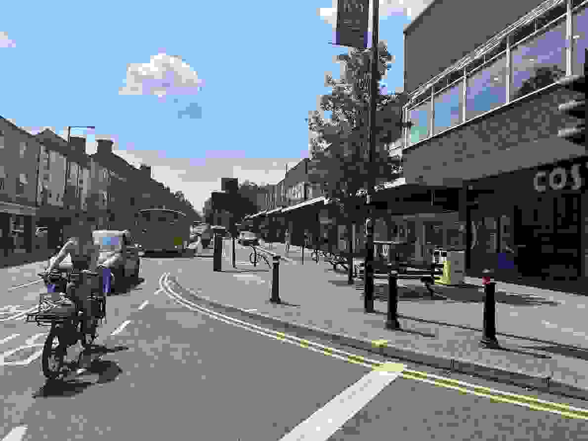 A cyclist cycles on the main road beside Broomhill Shopping Precinct in Sheffield. In the distance, parking spaces can be seen as well as trees and pedestrians. 