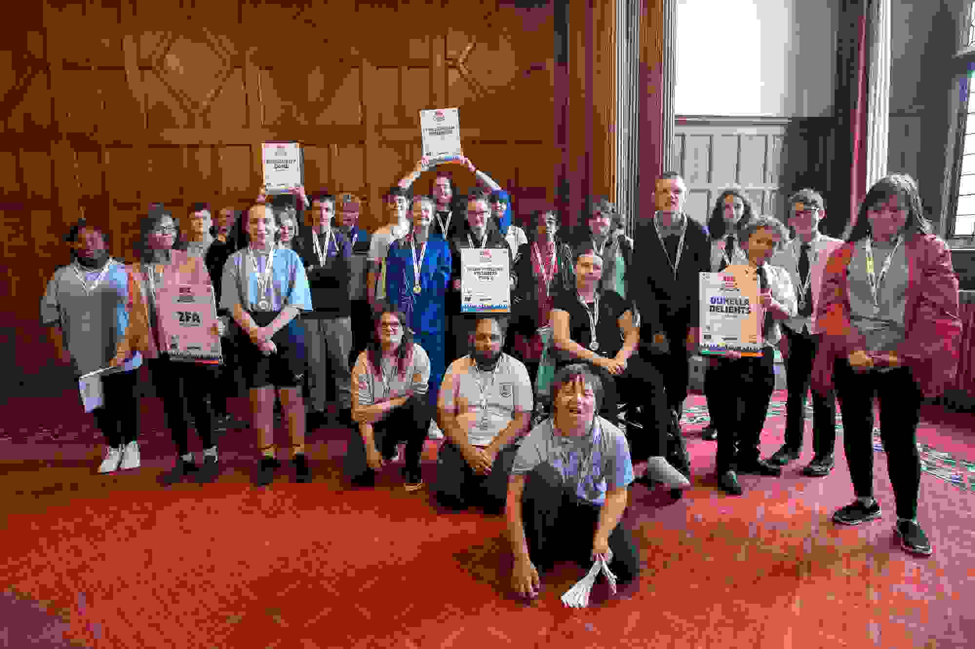 Group of students with their certificates and medal in the town hall with wooden panelling behind them and a red carpet