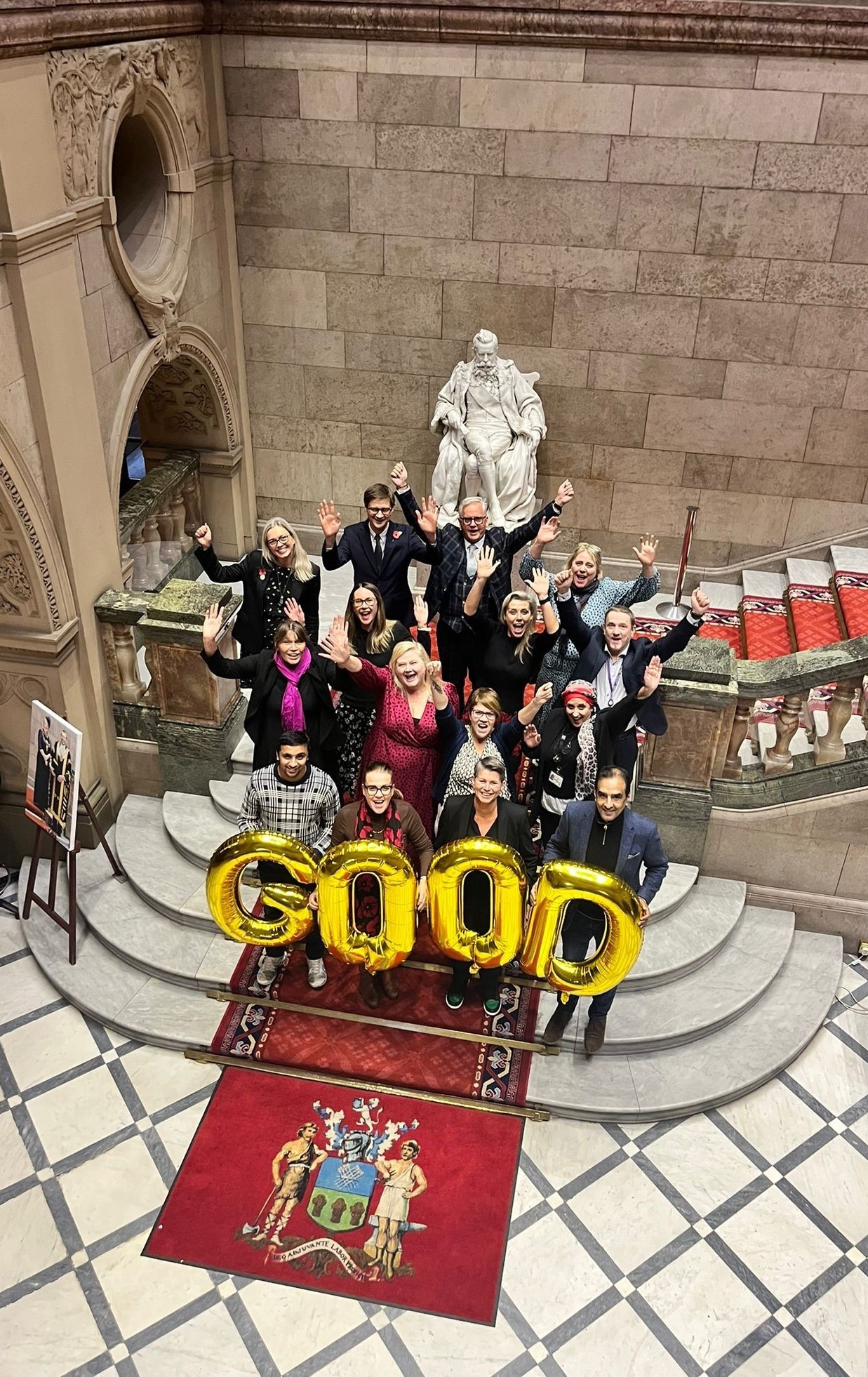 Officers and councillors stand on the bottom of the main staircase in Sheffield Town Hall with arms in the air celebrating the Ofsted result with four gold balloons held at the fron saying 'GOOD'