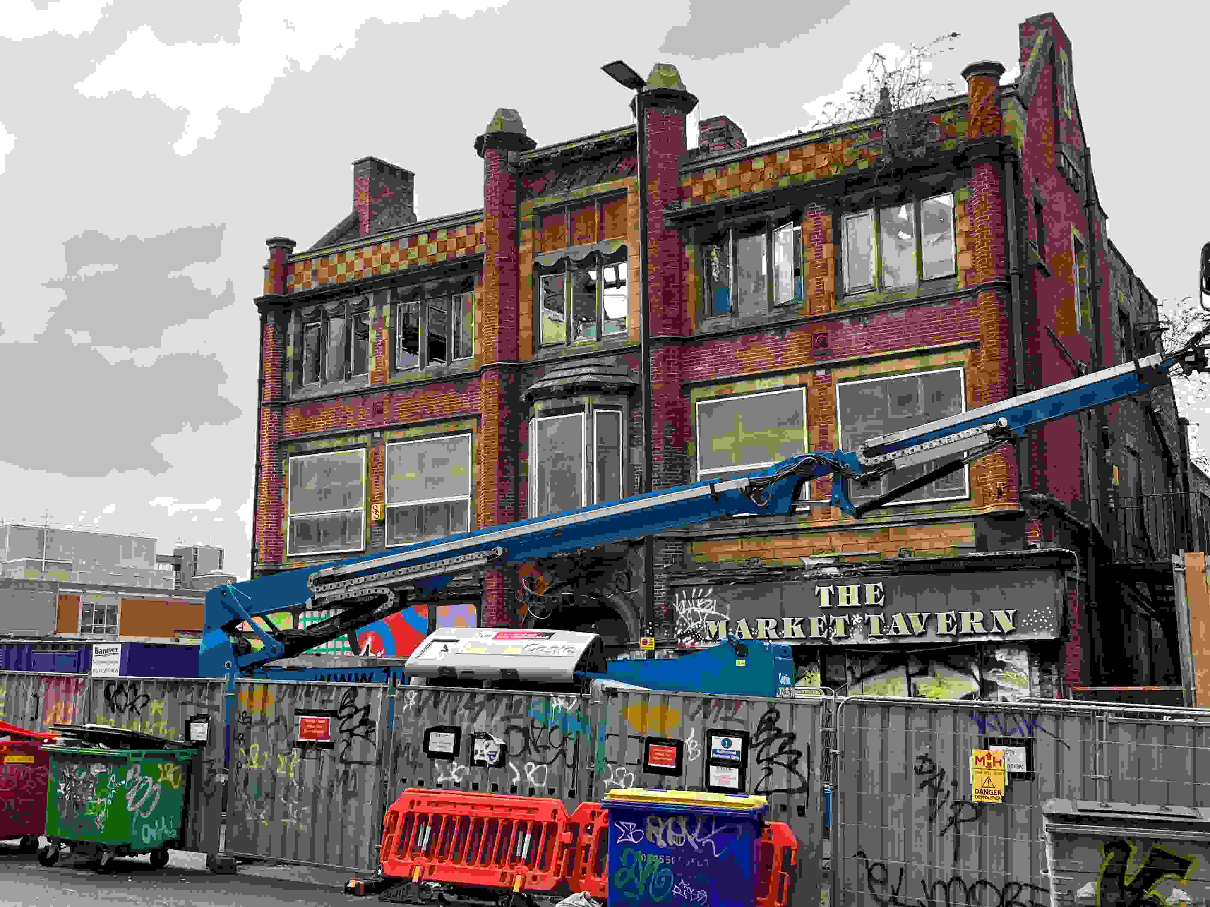The former Market Tavern pub house with demolition work evident on the upper floors with a blue cherry picker in front and the building is surrounded by hoardings