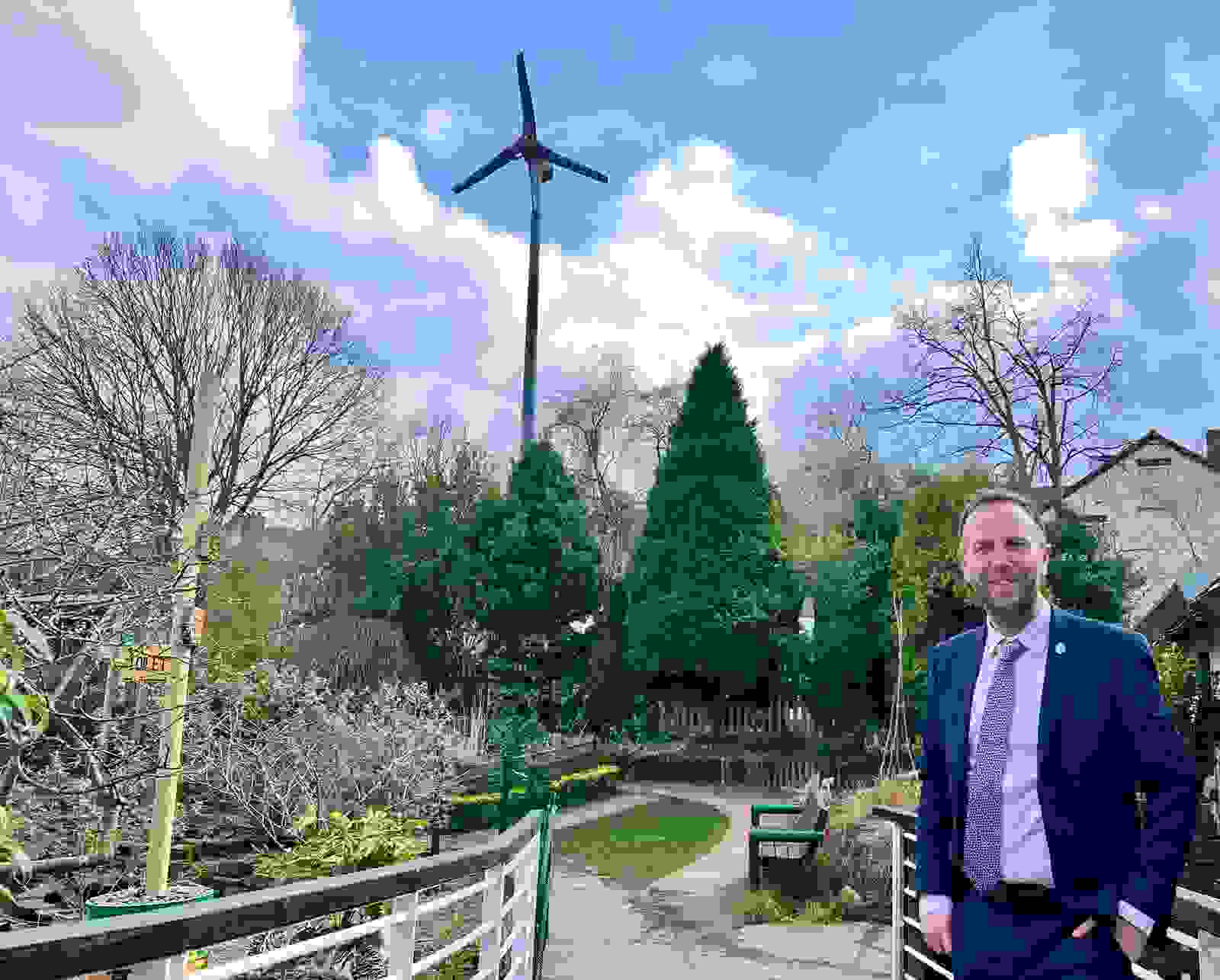 Cllr Ben Miskell stands in the foreground with a wind turbine in the background, scattered throughout the picture are a number of trees and a path disappears off into the distance