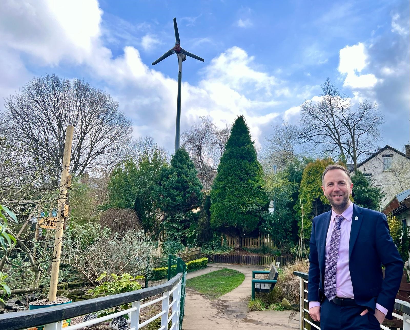 Cllr Ben Miskell stands in the foreground with a wind turbine in the background, scattered throughout the picture are a number of trees and a path disappears off into the distance