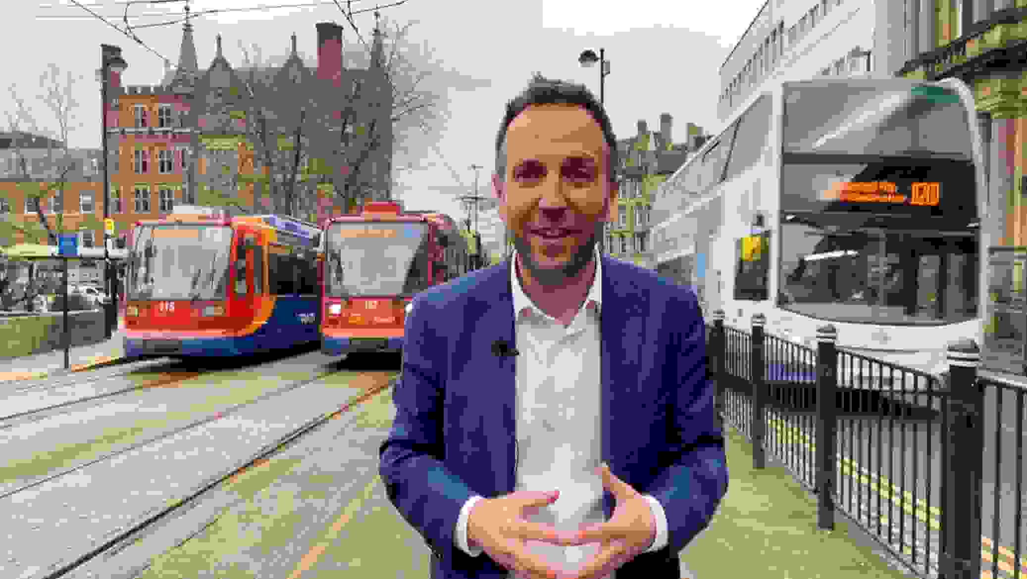 Cllr Ben Miskell stands in Sheffield city centre with two trams parked up behind him on his right hand side and a double decker bus on his left hand side
