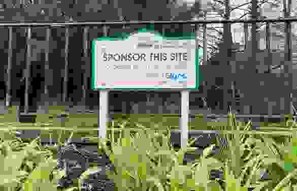 A blank, unused sponsorship sign stands among some grass in front of a metal railing with trees behind
