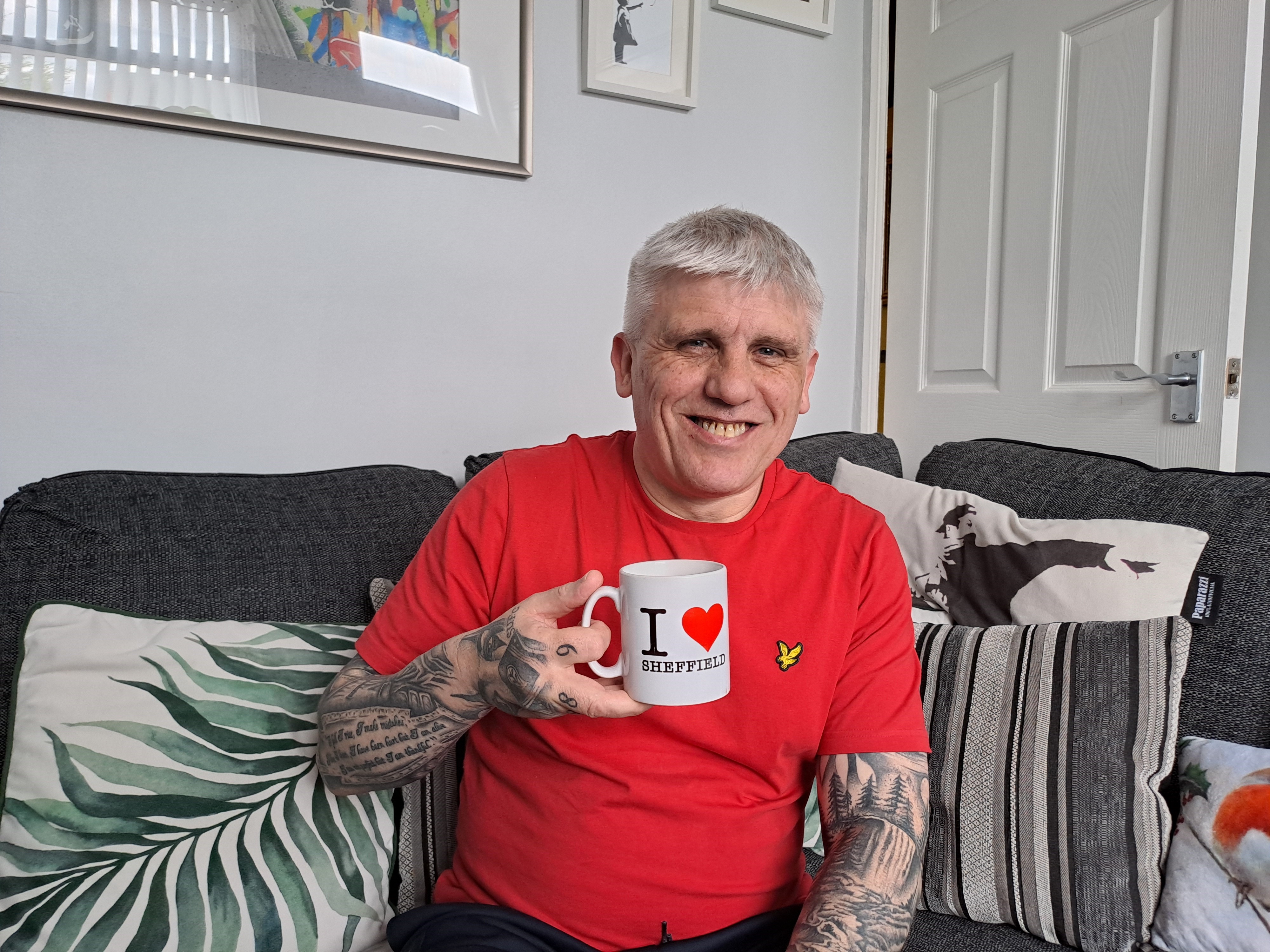 A smiling Mick Lewis enjoys a cup of tea on a sofa in a living room from a mug saying 'I Love Sheffield'