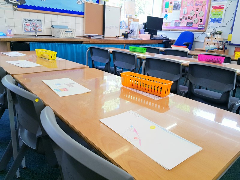 School classroom with light colour desks and dark blue chairs with artwork on the desks