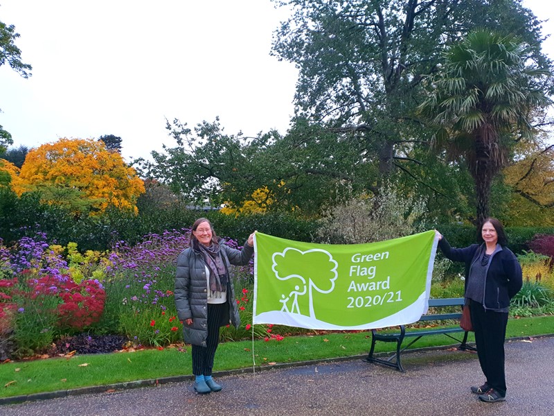 Cllr Mary Lea and Jill Thompson holding the green flag in the Sheffield Botanical Gardens with colourful plants in the background