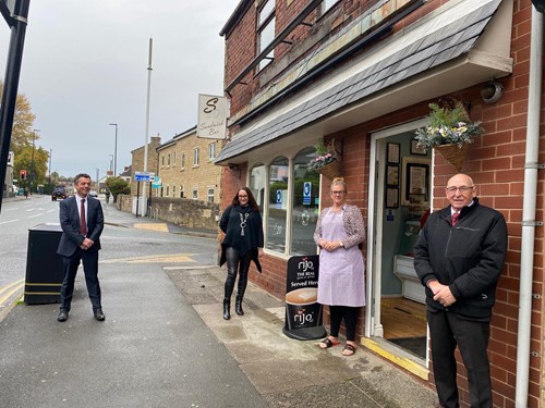 Four people stood socially distanced outside a shop front on a high street. A shop owner stands in an apron outside of the door.