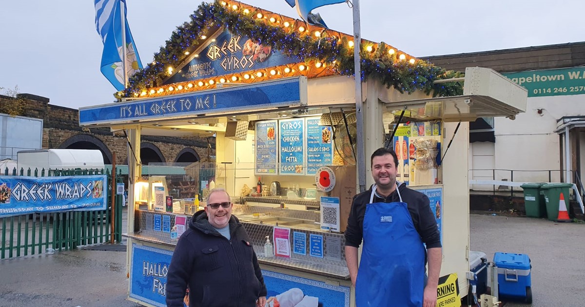 Two men stood in front a Greek food stand, one in an apron.