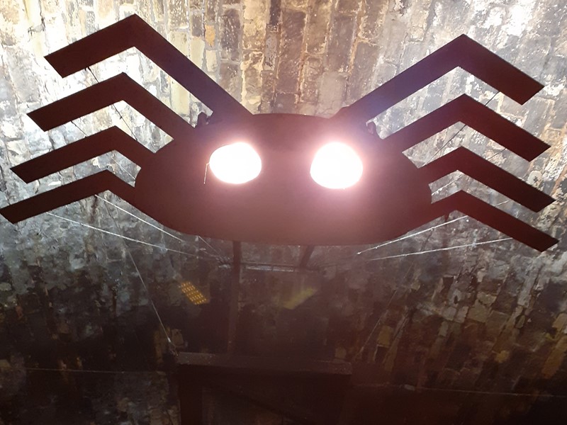 Dark metal spider hangs above bridge with white glowing eyes