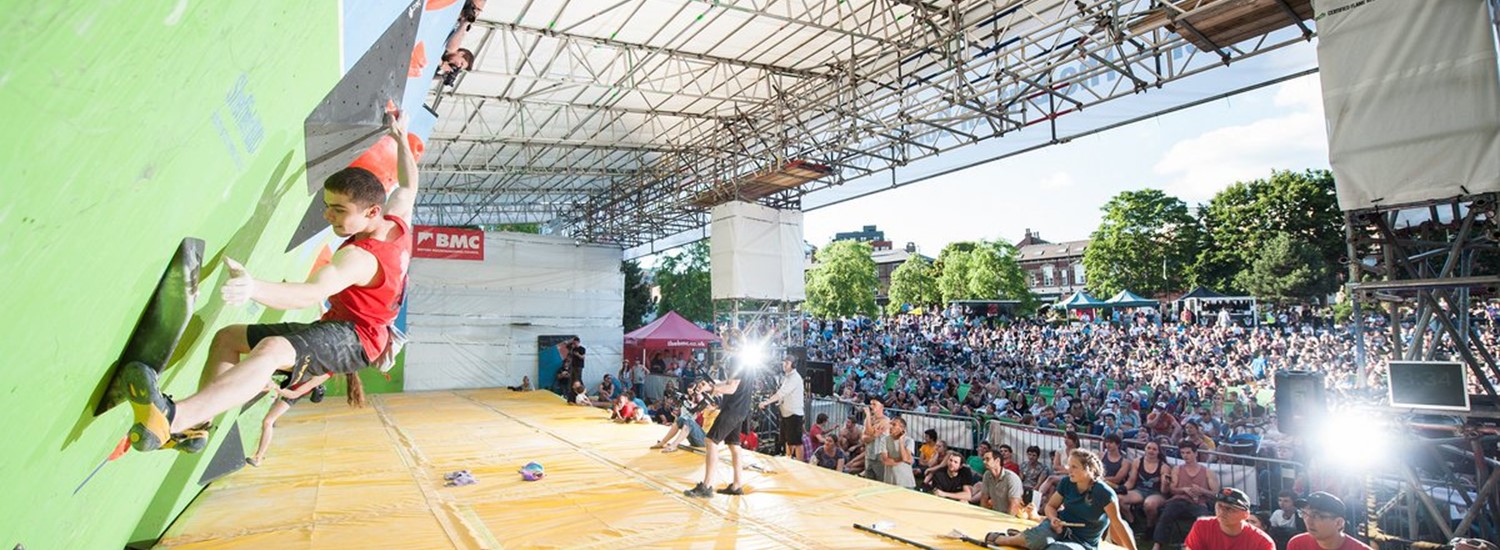 A man swings on a climbing wall with large crowd watching