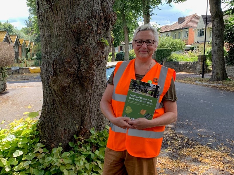 A woman in a high vis jacket stands in front of a tree on street