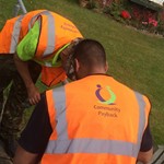 Two men wearing bright orange hi vis jackets which have 'Community Payback' written on them help repair a fence post