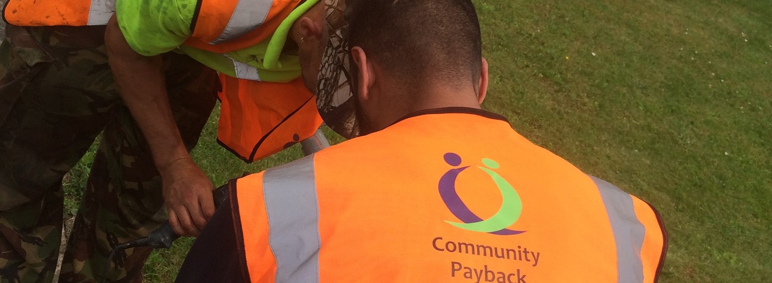 Two men wearing bright orange hi vis jackets which have 'Community Payback' written on them help repair a fence post