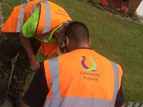 Two men wearing bright orange hi vis jackets which have 'Community Payback' written on them help repair a fence post