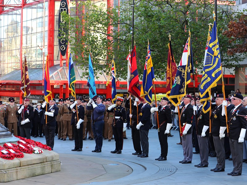 Men dressed in military uniform stand in a line holding flags, poppy wreaths lay in front of a monument