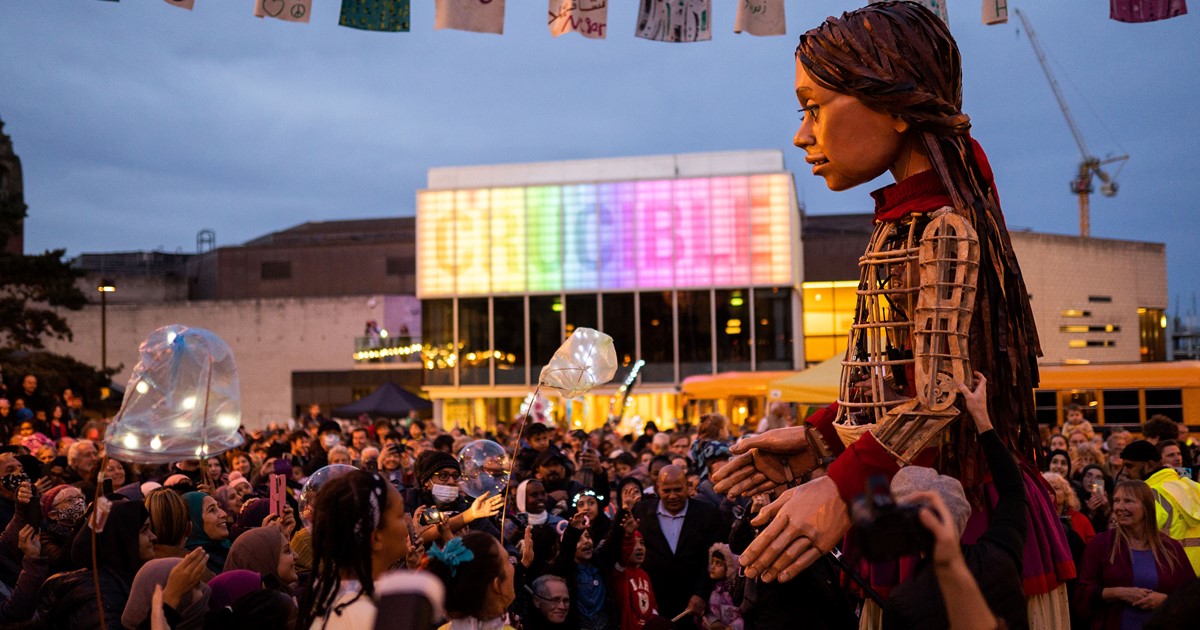 Little Amal, a 3.5metre high puppet of a Syrian refugee, dances with people in Sheffield outside the Crucible which is lit up with rainbow lights