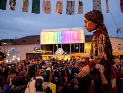 Little Amal, a 3.5metre high puppet of a Syrian refugee, dances with people in Sheffield outside the Crucible which is lit up with rainbow lights