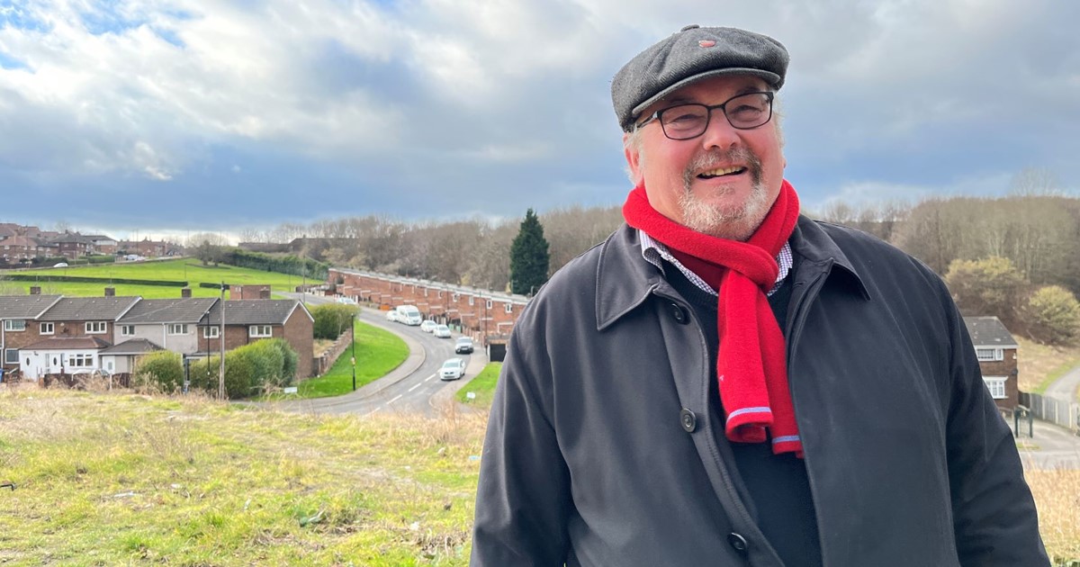 Cllr Terry Fox at Corker Bottoms with green space and housing around him with a blue sky and white fluffy clouds. Cllr Fox is wearing a grey overcoat, red scarf and a wool cap