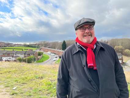 Cllr Terry Fox at Corker Bottoms with green space and housing around him with a blue sky and white fluffy clouds. Cllr Fox is wearing a grey overcoat, red scarf and a wool cap