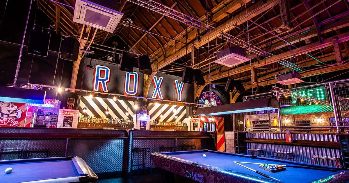 Two pool tables with a bar behind and a neon sign spelling Roxy