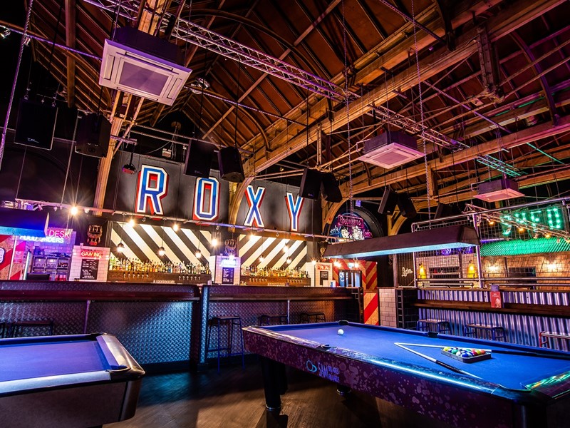 Two pool tables with a bar behind and a neon sign spelling Roxy
