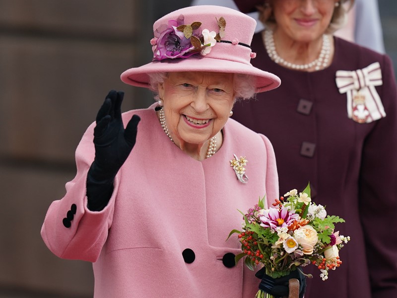 Her Majesty the Queen in a pink coat and hat waving with a black glove on her hand and holding a floral post in the other