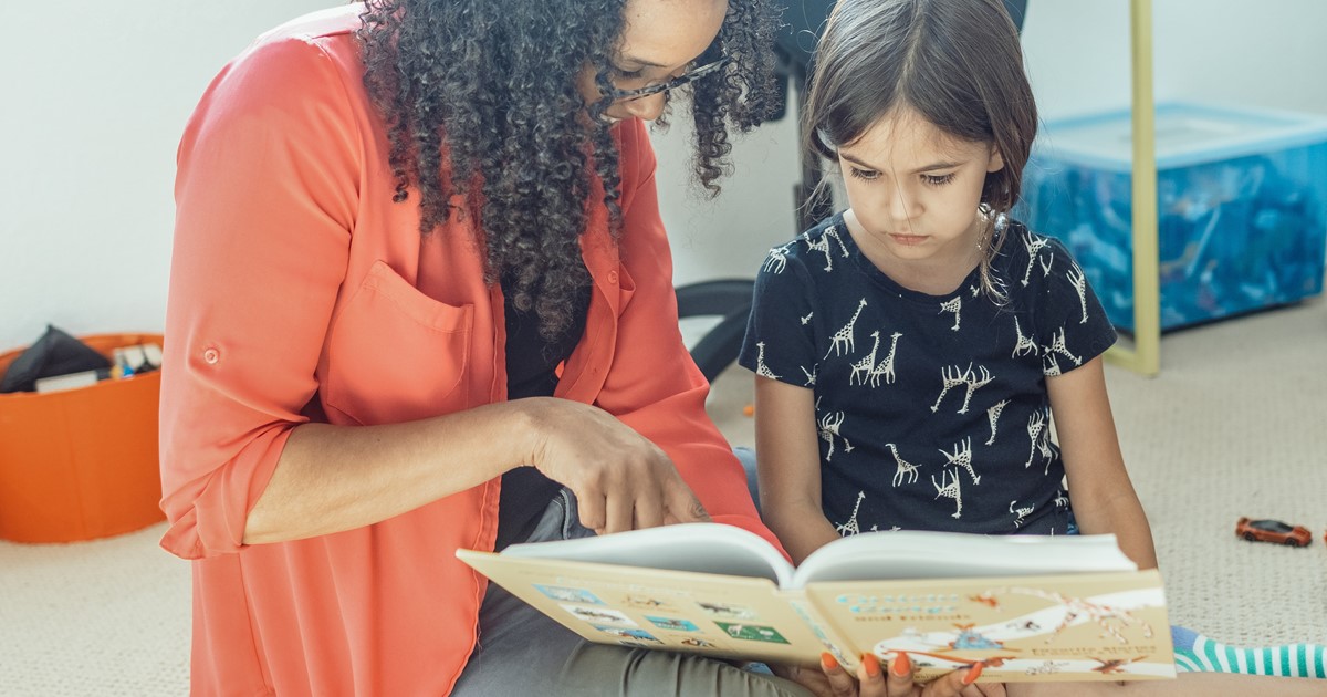A woman in a red shirt is reading a children's book with a little girl