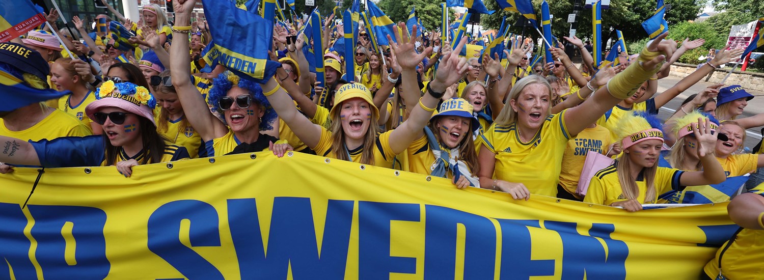 A large group of people dressed in yellow hold a large banner that say 'camp Sweden'