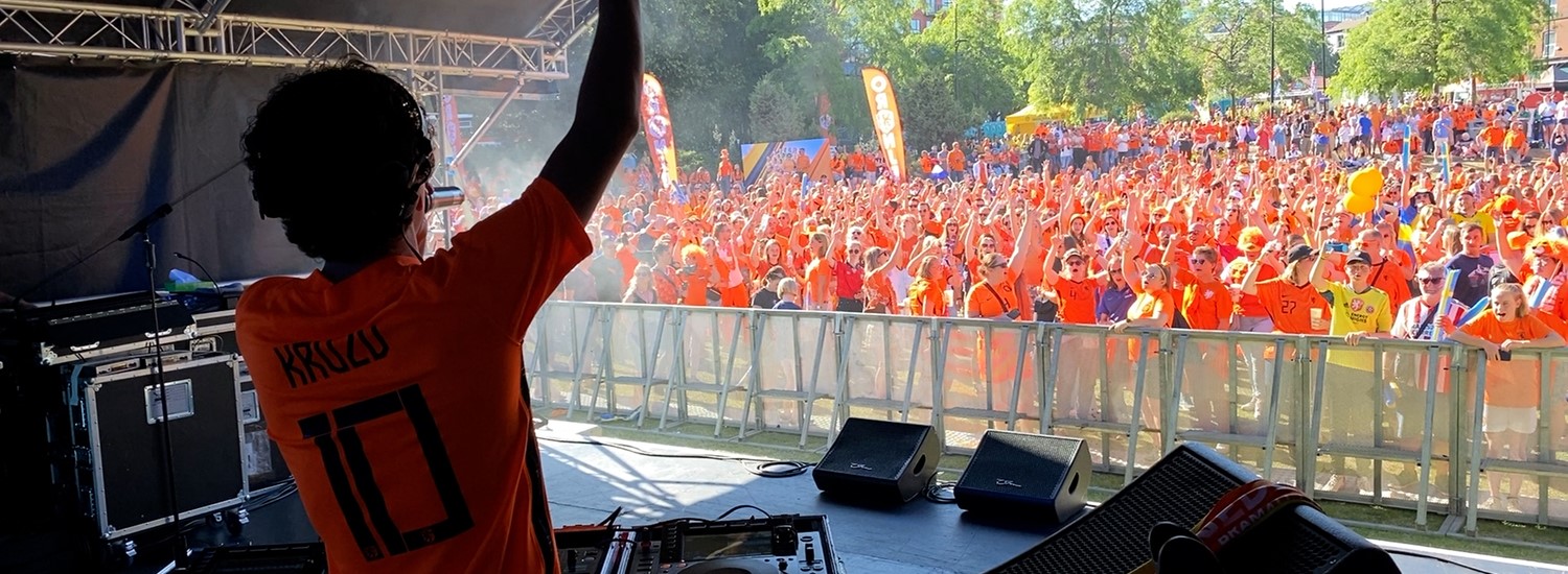 A man stands behind dj decks on a stage, a large crowd of people stand in front of the stage