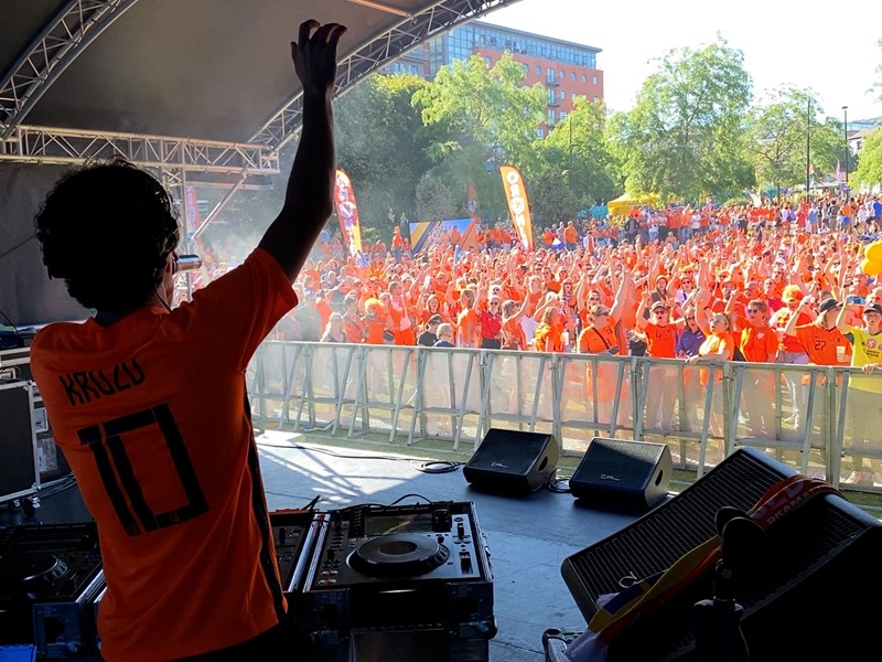 A man stands behind dj decks on a stage, a large crowd of people stand in front of the stage
