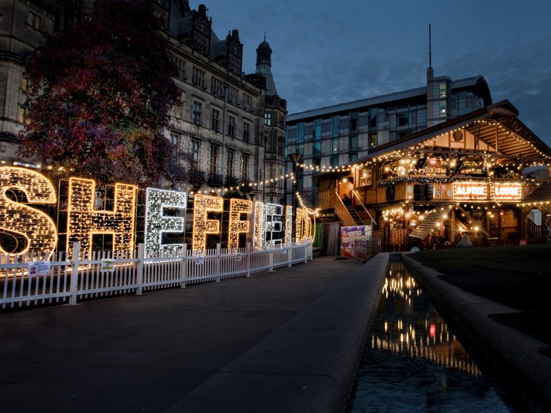 The word 'Sheffield' in gold and silver lights, a large civic building is behind it and a wooden festive bar