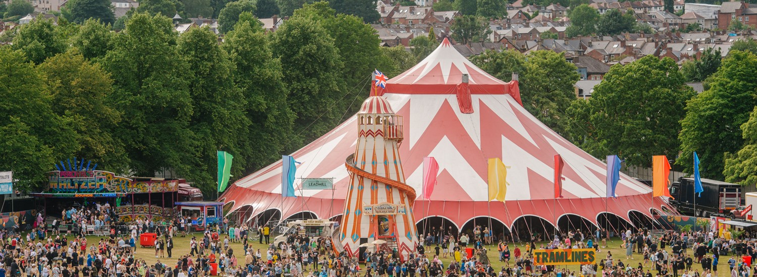 A huge crowd of people stand around a large festival tent, trees are in the background