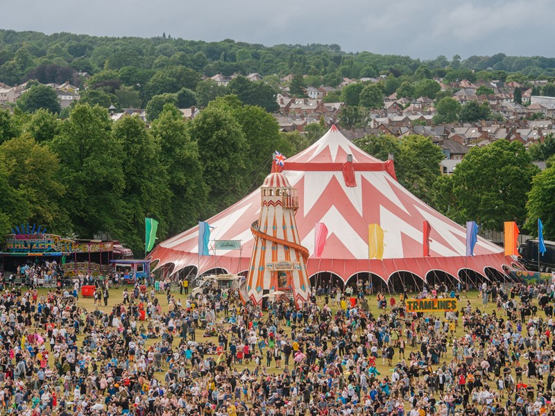 A huge crowd of people stand around a large festival tent, trees are in the background