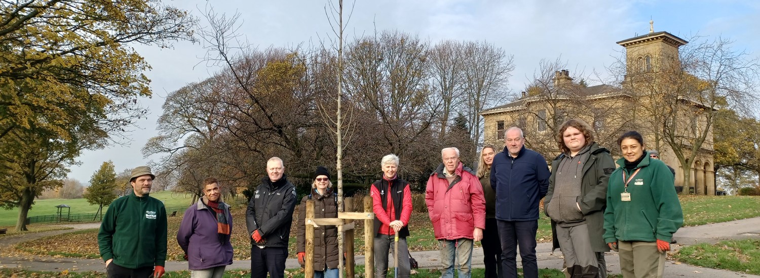 A group of people stand beside a newly planted elm tree in a park a large stone building is the background