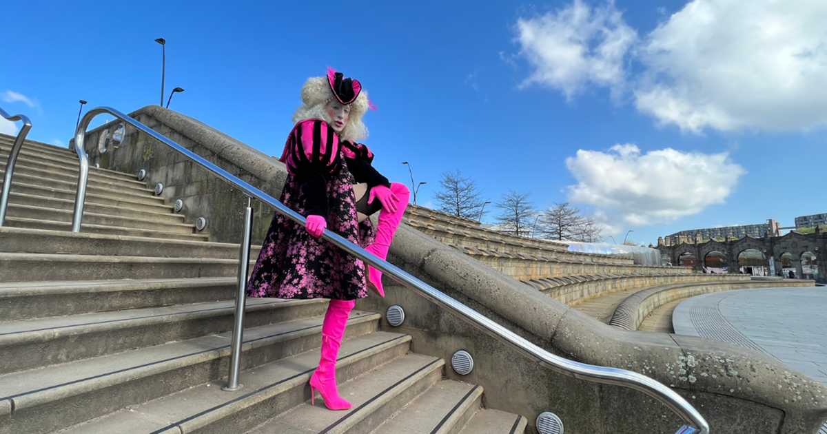 Jamie is wearing a pink and white outfit and a white curly wig. He is standing on some steps looking down at the camera and posing.