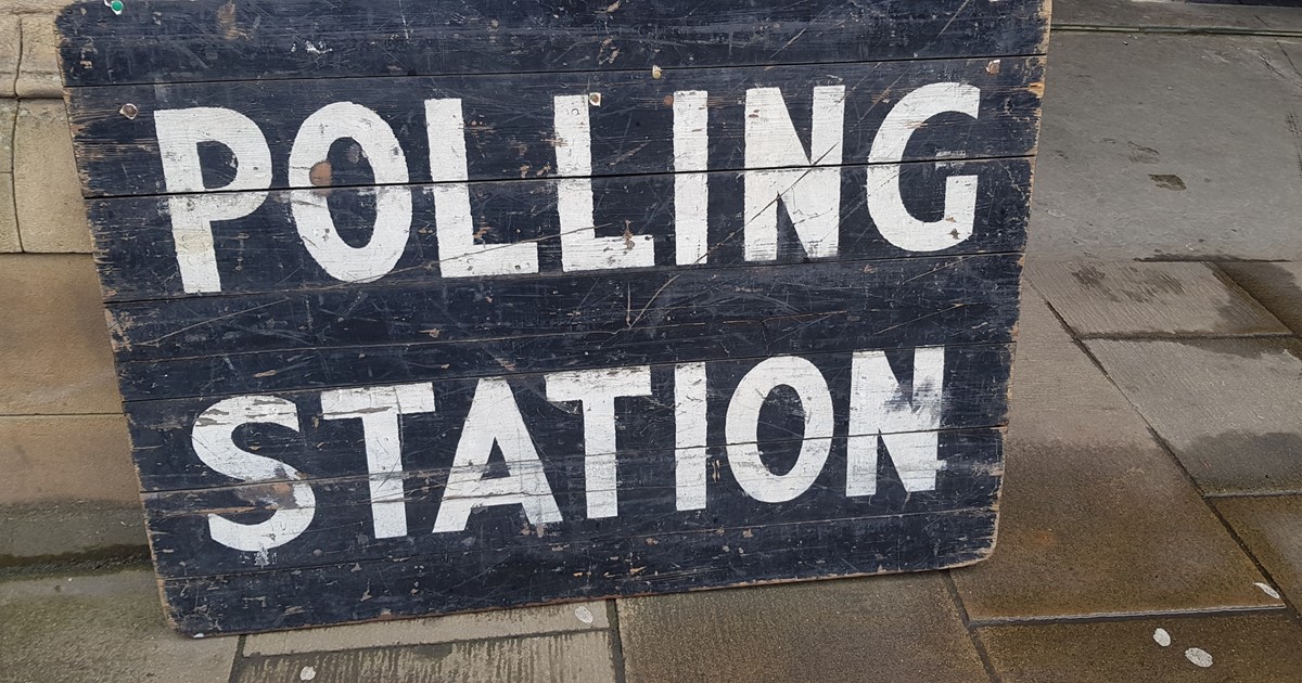 A polling station sign containing white text saying 'Polling Station' on a black background, the sign is leaning against a brick wall