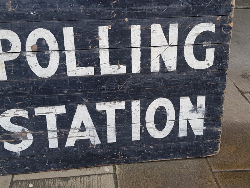 A polling station sign containing white text saying 'Polling Station' on a black background, the sign is leaning against a brick wall
