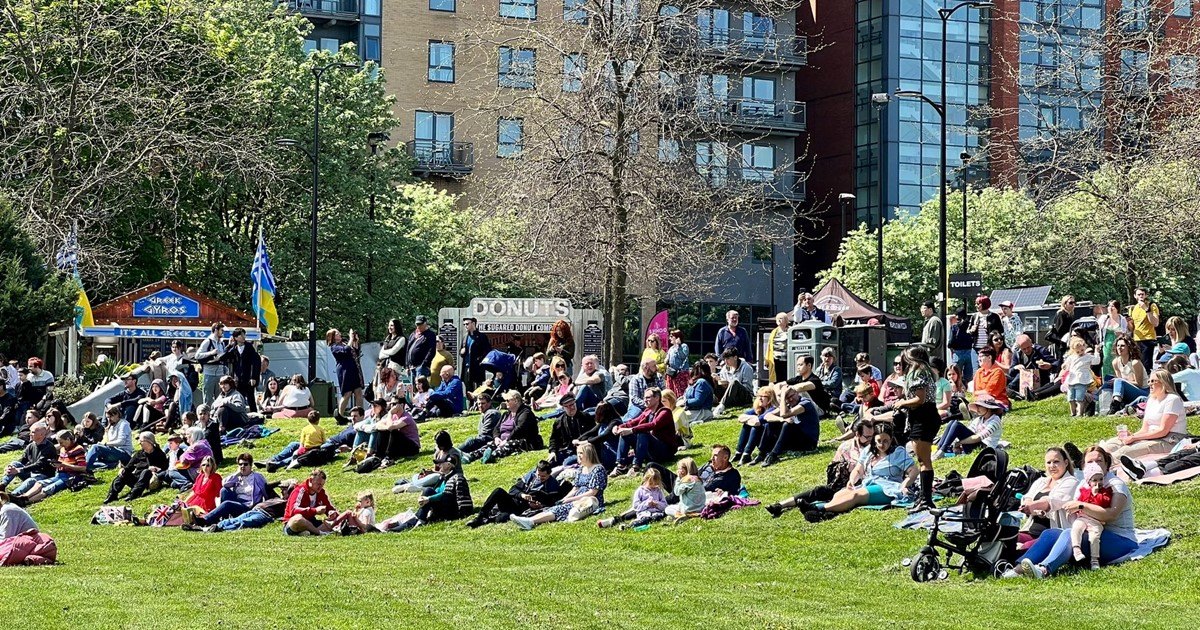 People sit on Devonshire Green in the sunshine watching acts perform on stage.