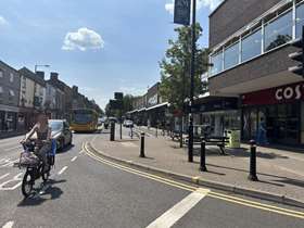 A cyclist cycles on the main road beside Broomhill Shopping Precinct in Sheffield. In the distance, parking spaces can be seen as well as trees and pedestrians. 