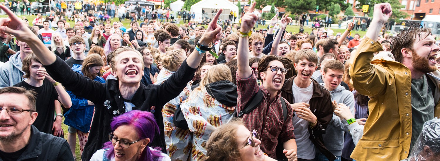 A crowd of people facing the camera with their hands in the air whilst they watch a band