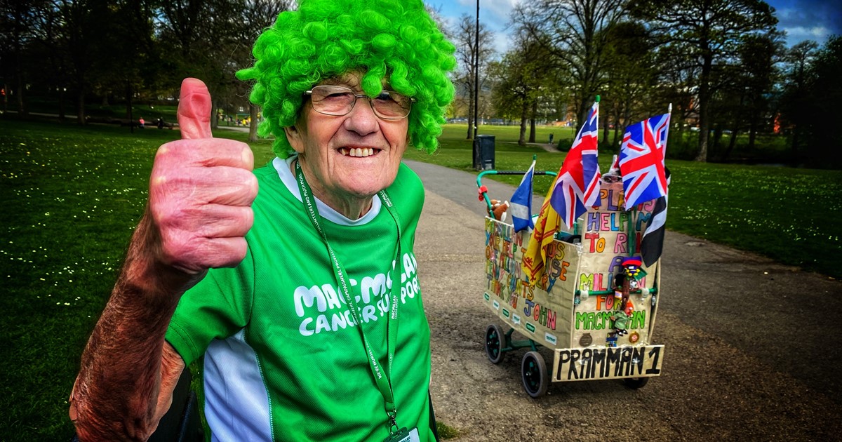 John Burkhill smiling, in a Macmillan t-shirt and green wig, with his thumb up at the camera. His famous 'pram' is seen to the right of him.