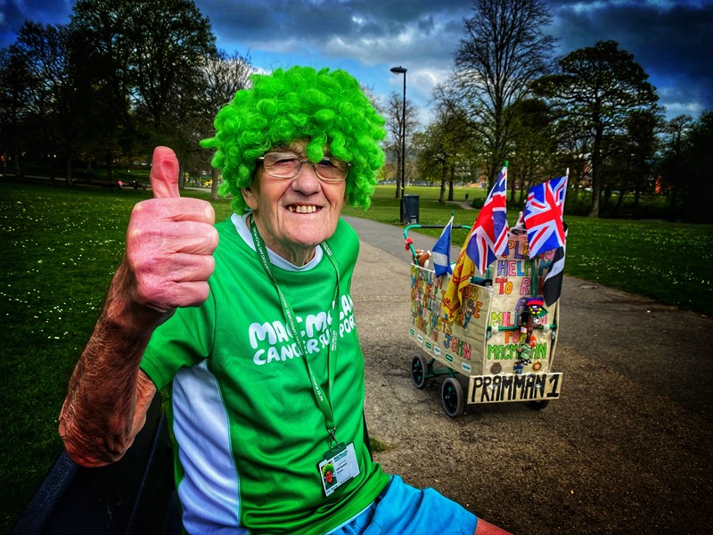 John Burkhill smiling, in a Macmillan t-shirt and green wig, with his thumb up at the camera. His famous 'pram' is seen to the right of him.