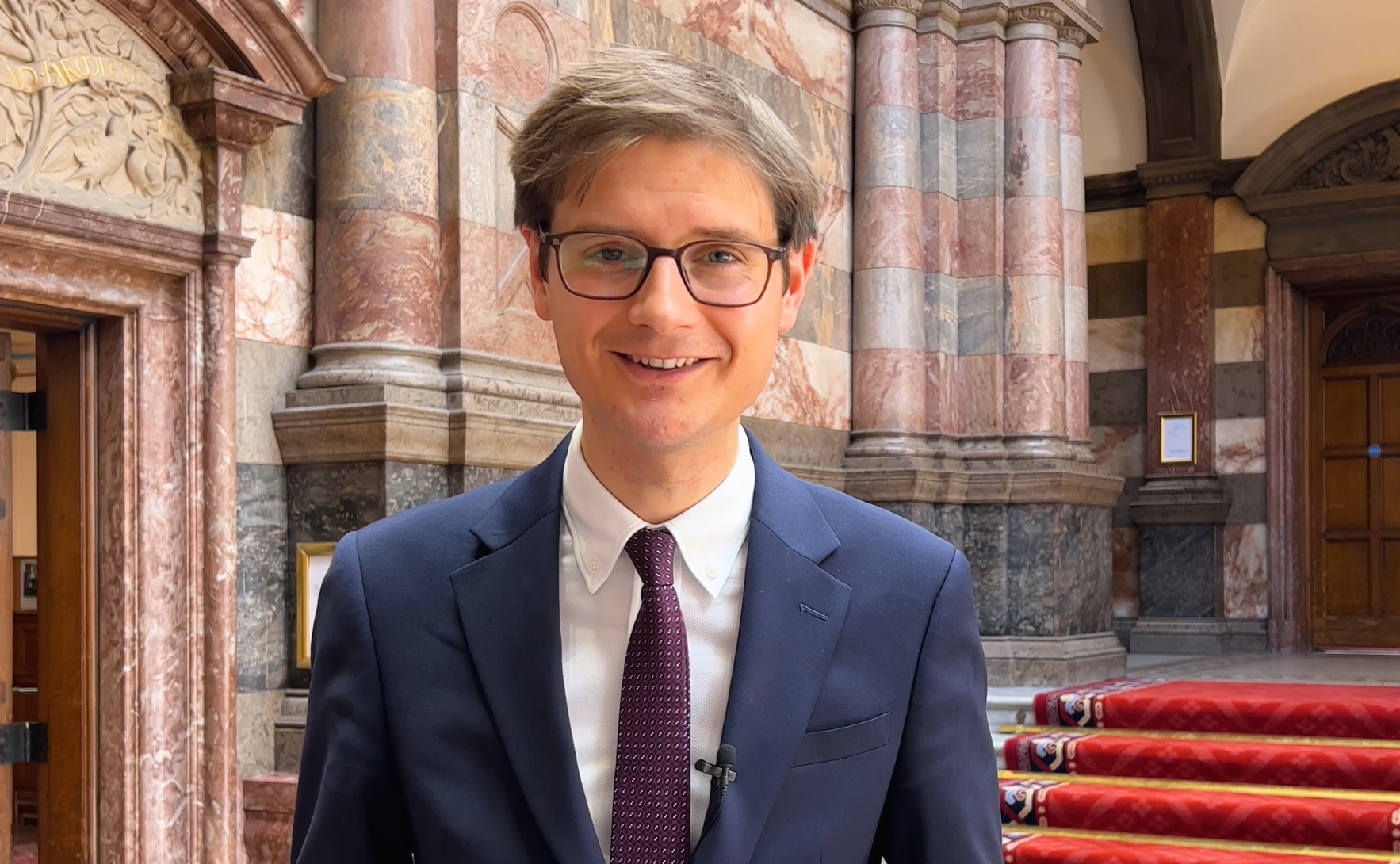 Councillor Tom Hunt, wearing a blue suit and glasses, standing in the Town Hall smiling 